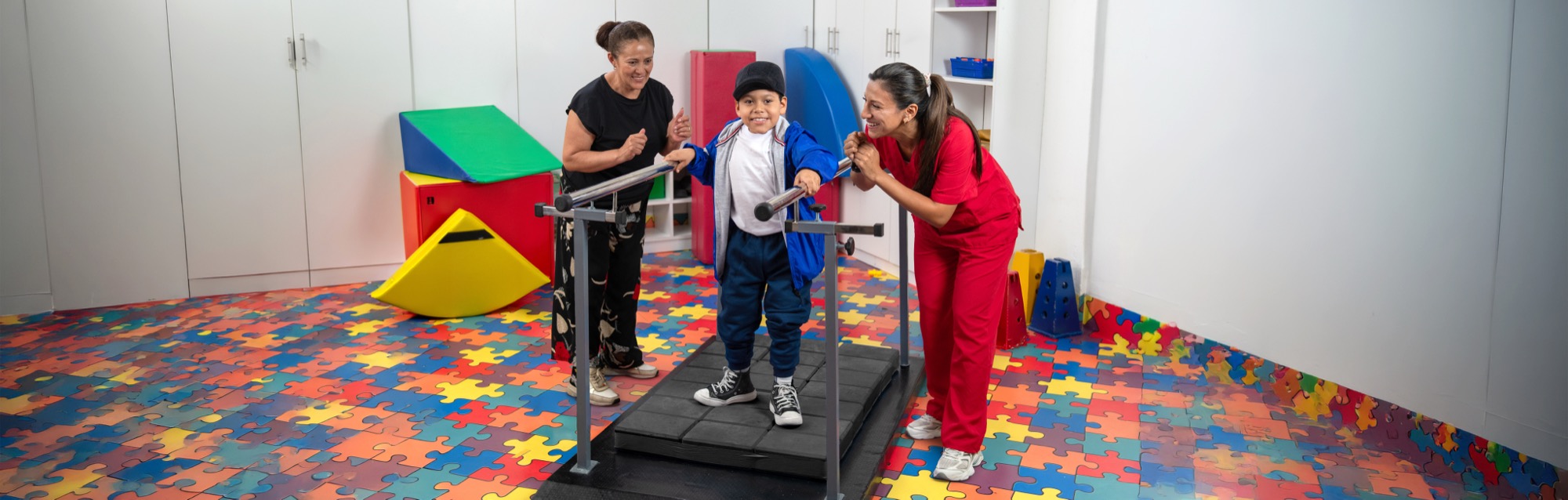 Un niño camina entre barras paralelas durante una sesión de terapia de rehabilitación, acompañado por dos terapeutas.